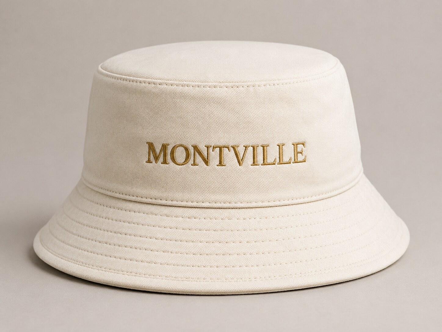 A cream cotton Montville bucket hat with the wordmark embroidered in gold.