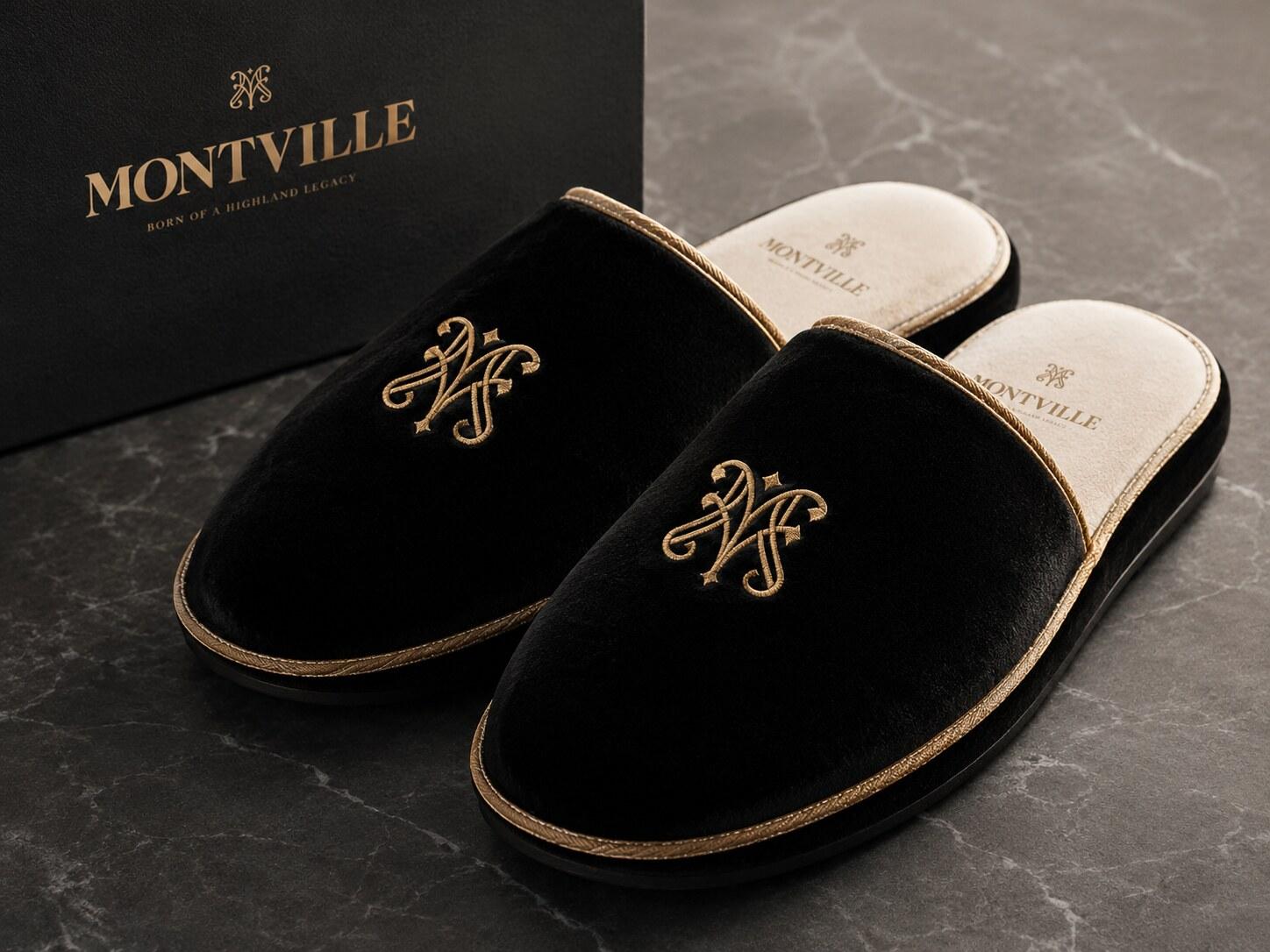 A pair of black velvet Montville slippers with the gold monogram, on marble beside their black presentation box.